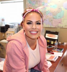 a picture of Sunshine Ray wearing a pink hoodie, white shirt, and pink headband in an office setting